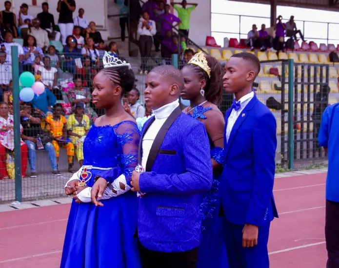 Students in royal blue gowns and suits, two wearing crowns, at a school celebration in a stadium